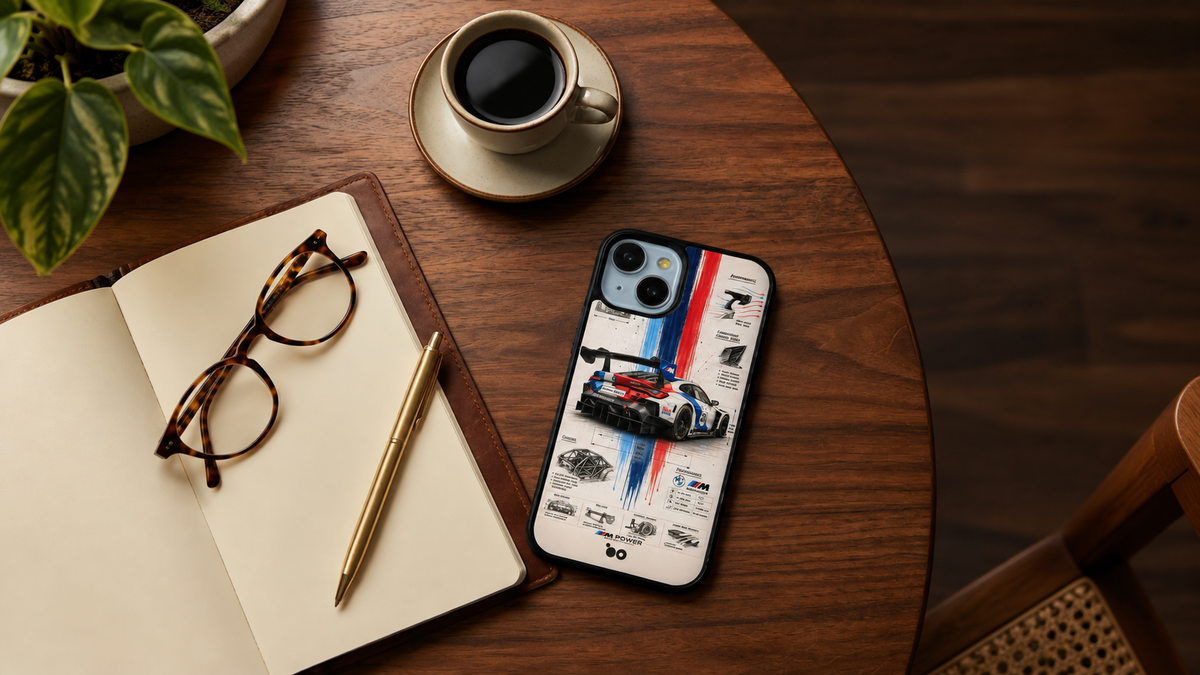 A smartphone case featuring a racing car graphic and the BMW logo, displayed on a round wooden table next to an open notebook, eyeglasses, a pen, and a cup of coffee.